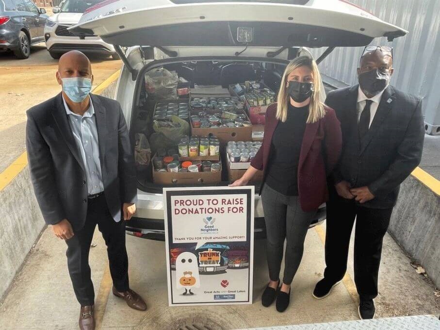 Three masked people by an open trunk filled with canned goods from Good Neighbors Food Donation event