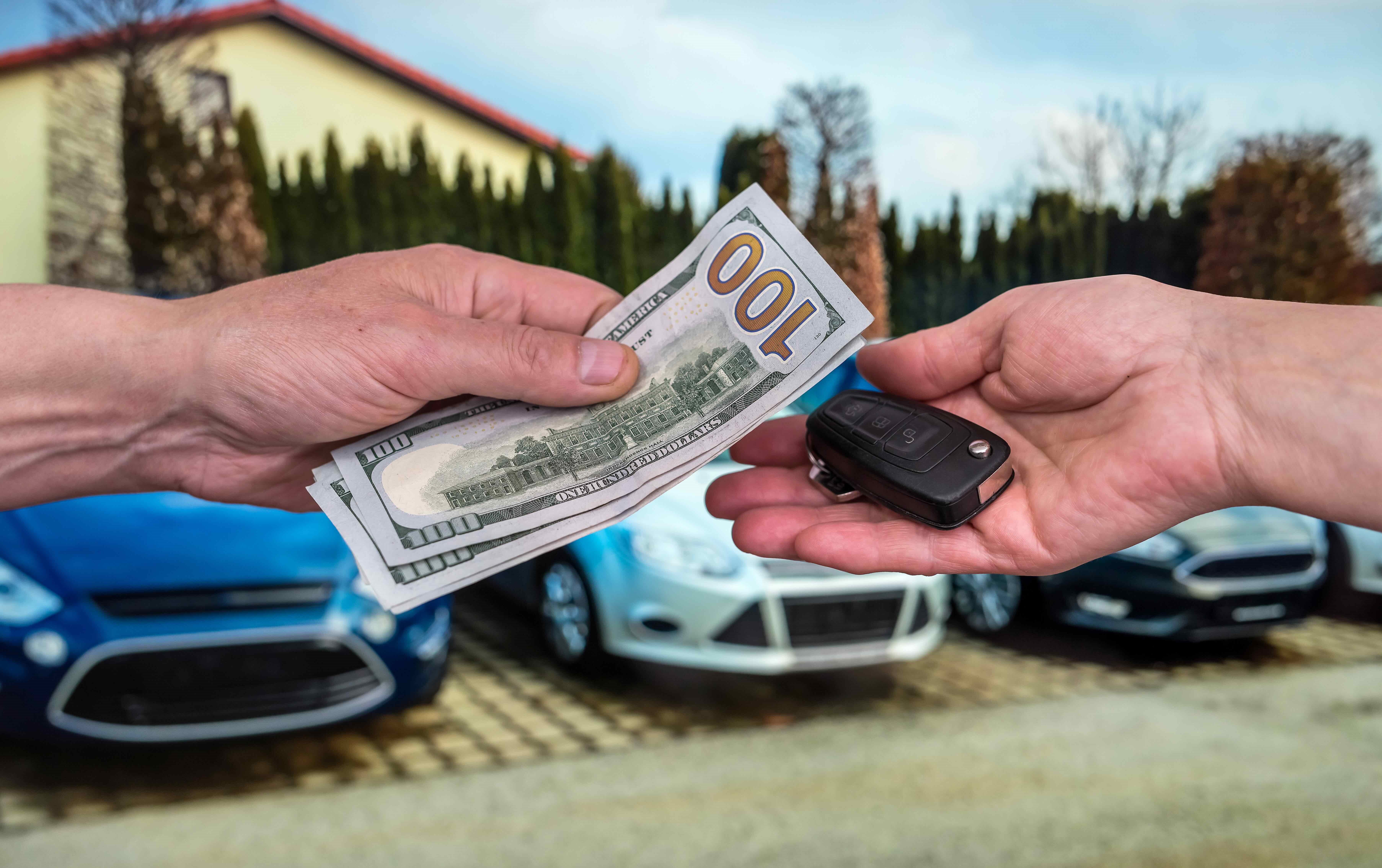Hands exchanging cash and car keys in front of cars
