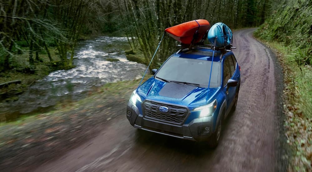 A blue 2022 Subaru Forester Wilderness is shown with kayaks on the roof driving by a river.