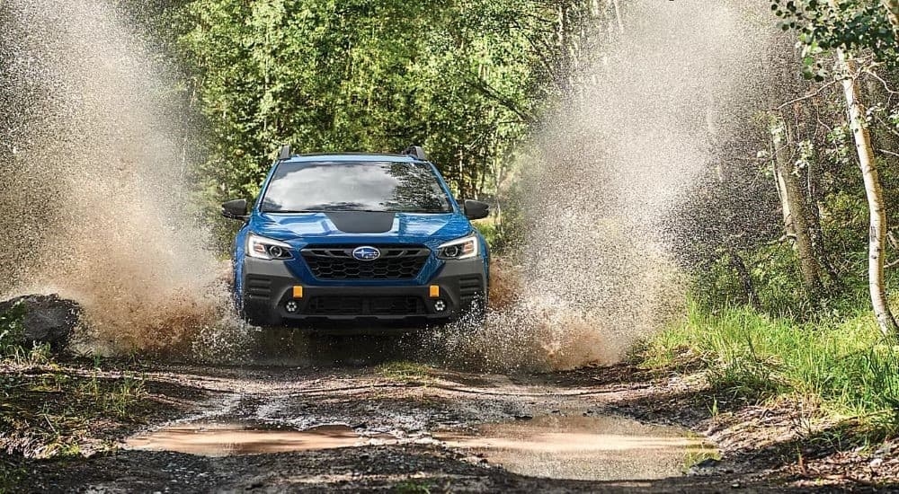 A blue 2023 Subaru Outback Wilderness is shown splashing through a puddle in the woods.