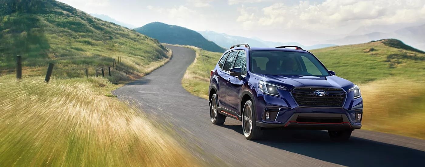 A blue 2023 Subaru Forester is shown driving on a road through a winding, hilly field.