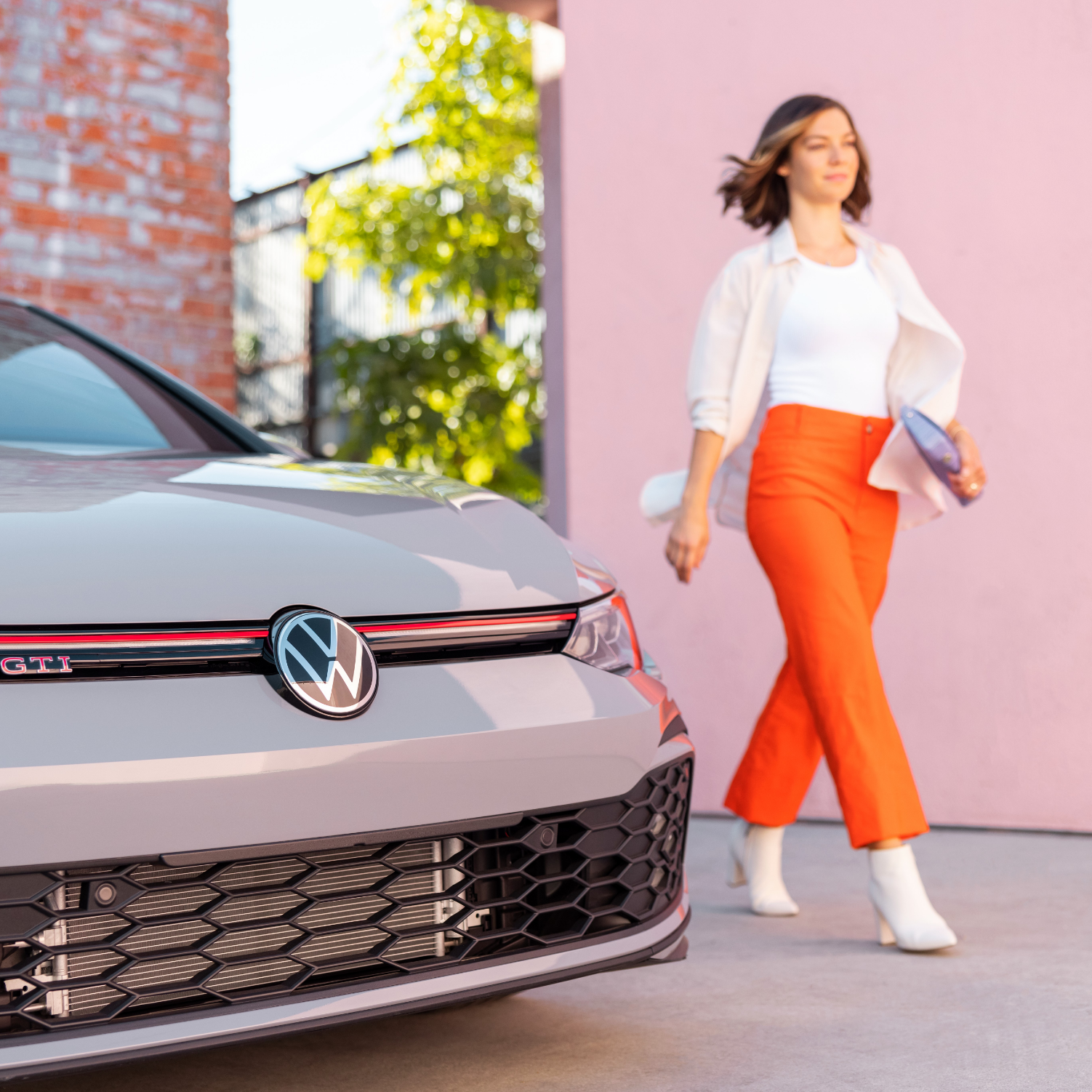 Woman in bright orange pants walking past front of VW sedan.