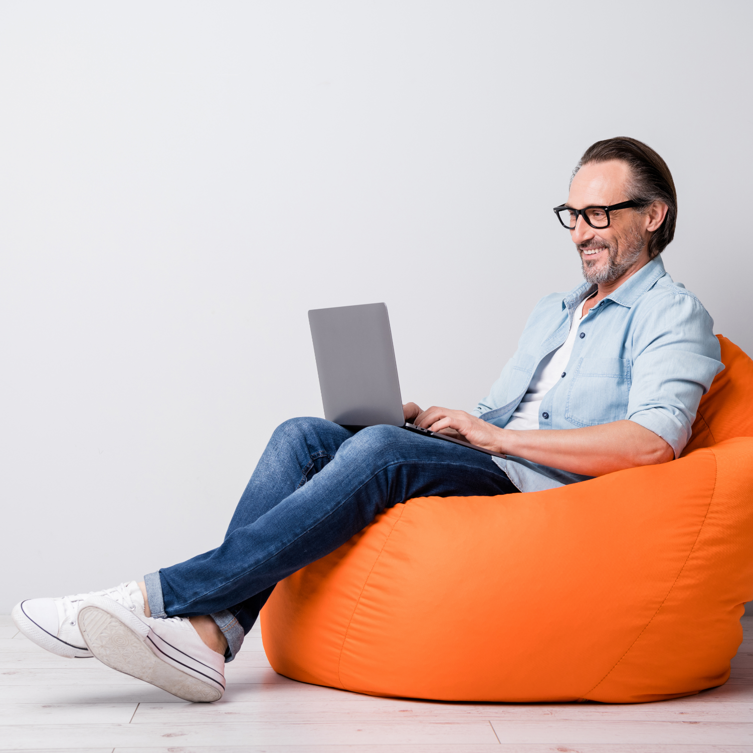 Smiling man sitting on orange beanbag working on his laptop computer.