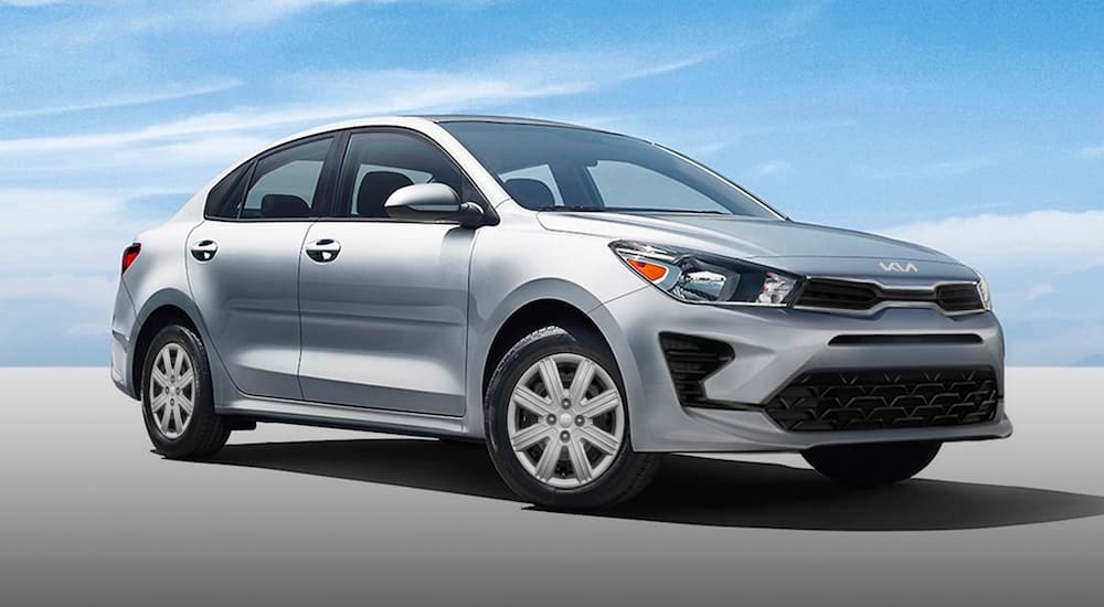 A silver 2023 Kia Rio parked on pavement against a blue sky with clouds.