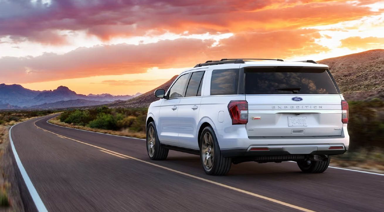 White Ford Expedition SUV driving towards sunset lit sky on desert road with mountains in the background.