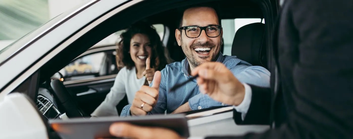 A couple is shown smiling from the front seat of a car.