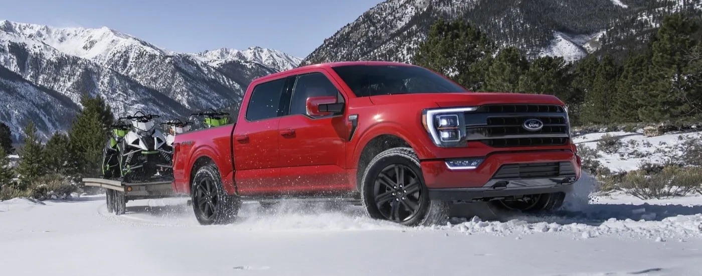A red 2021 used Ford F-150 for sale is shown towing snowmobiles through the snow.