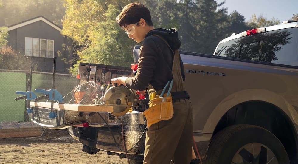 A person is shown using a circular saw on lumber attached to the bed of a grey 2022 Ford F-150 Lightning.