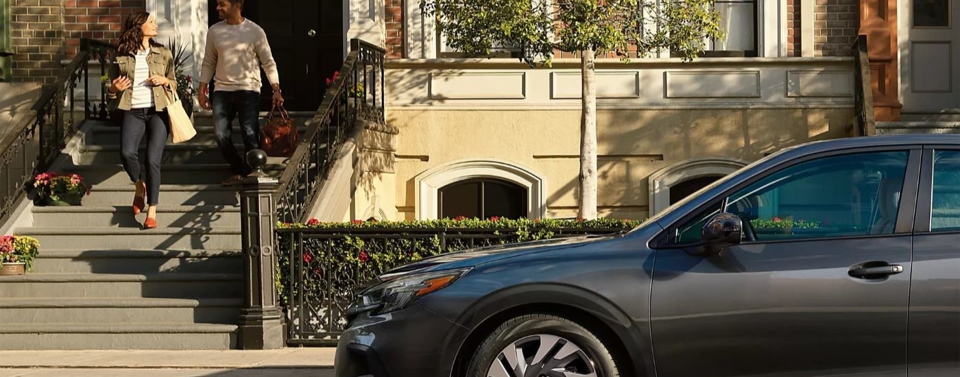 A dark grey 2024 Subaru Legacy Limited is shown near a couple walking down a set of stairs.