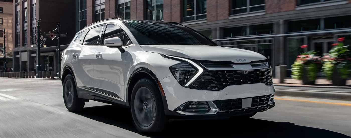 A white 2023 Kia Sportage Hybrid is shown driving on a city street lined with tall brick buildings.