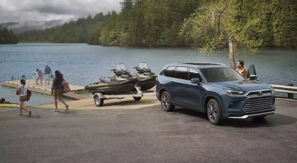 A grey 2024 Toyota Grand Highlander is shown towing jet skis near a lake.