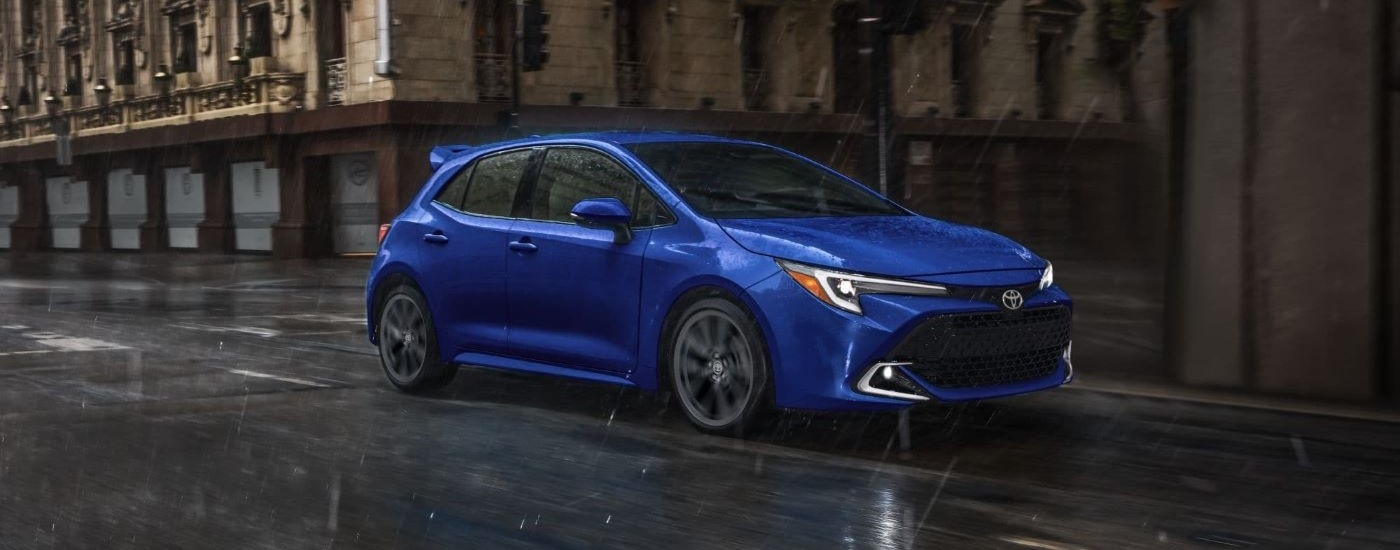A blue 2024 Toyota Corolla is shown driving on a city street in the rain.
