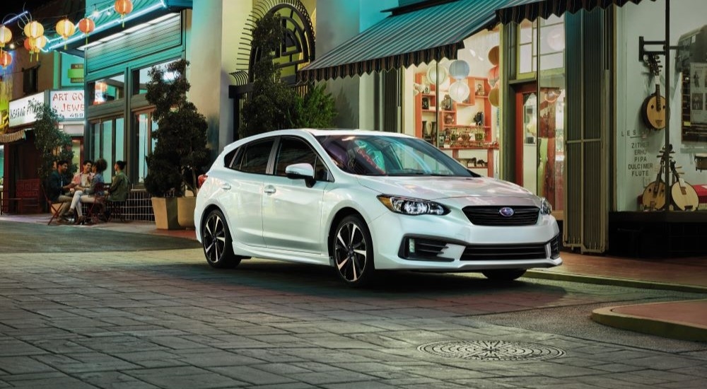 A white 2021 Subaru Impreza is shown parked in front of a row of shops.