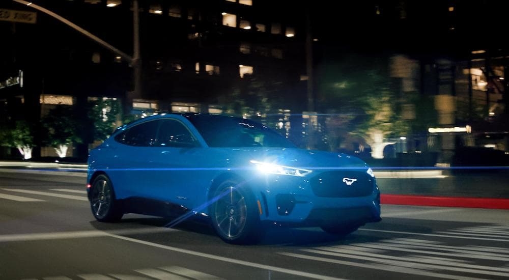 A blue 2024 Ford Mustang Mach-E for sale is shown from the front at an angle on a city street.