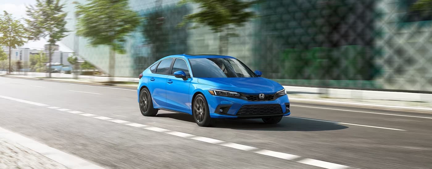 A blue 2024 Honda Civic Hatchback Sport Touring driving on a street near a Honda dealer.