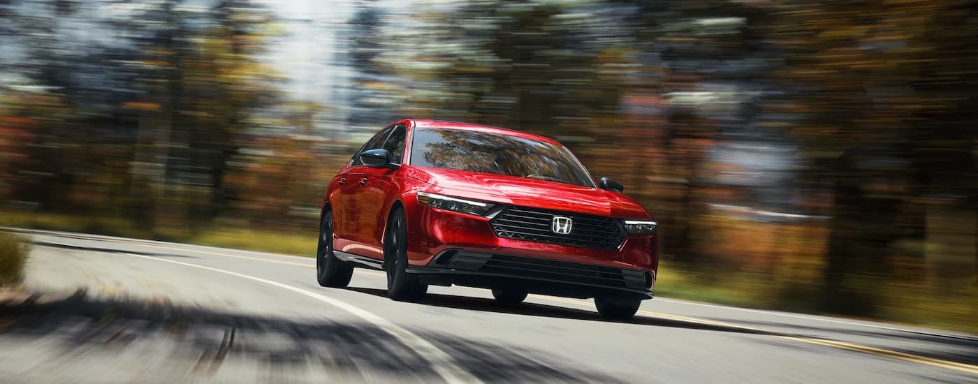 A red 2023 Honda Accord Sport-L driving on a winding road lined with colorful trees.