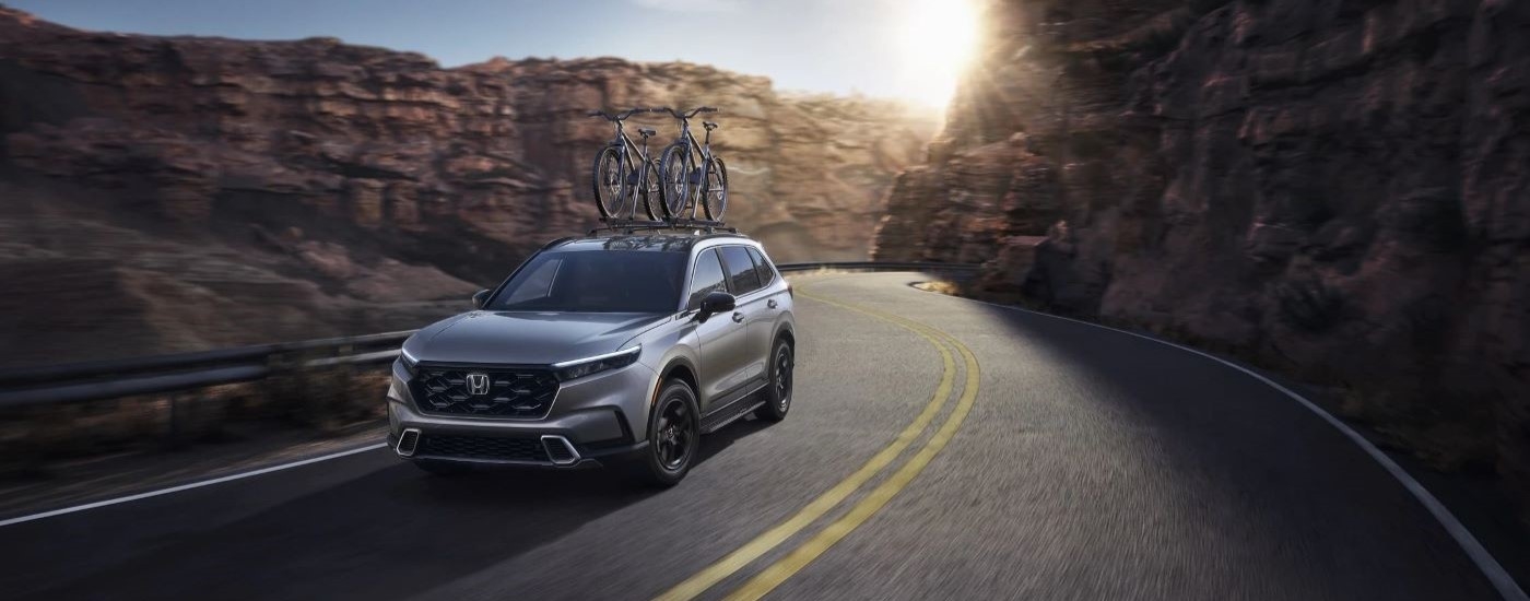 A grey 2023 Honda CR-V Hybrid with bicycles on the roof rack driving on mountain road.