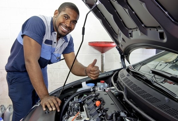 Smiling mechanic giving a thumbs-up while working on a Volkswagen car engine in an auto repair shop.