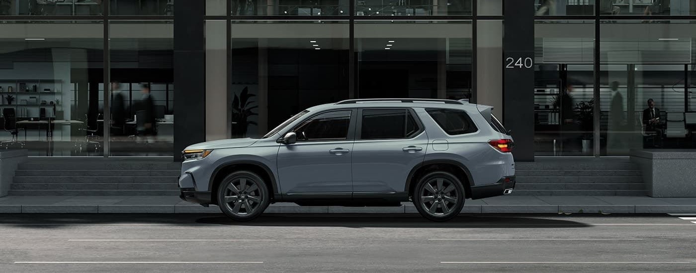 A grey 2023 Honda Pilot is shown from the side driving on a city street in front of a glass building.