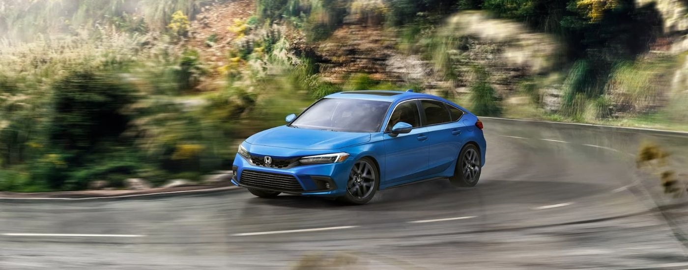 A blue 2024 Honda Civic Sport Touring is shown driving around a bend on a road.