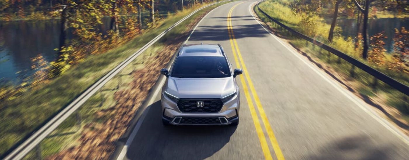 A popular used Honda CR-V for sale, a silver 2023 Honda CR-V, is shown driving past a lake.