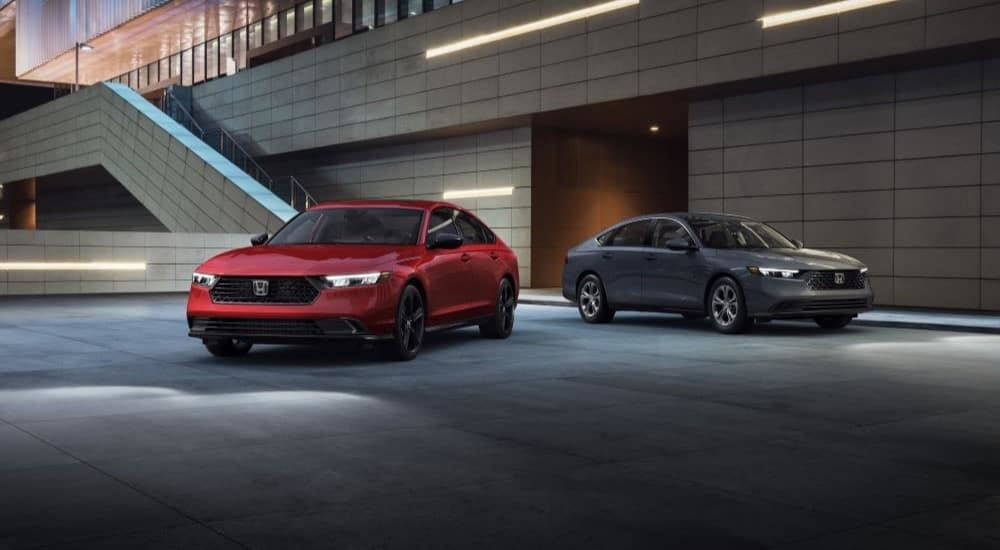 Two used hybrid vehicles for sale, one red and one grey 2023 Honda Accord Sport LX Hybrid's, are shown parked in a garage.