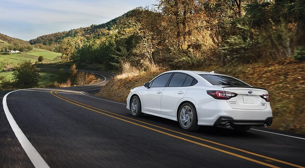 Rear view of a white 2025 Subaru Legacy Sport driving on a winding country road.