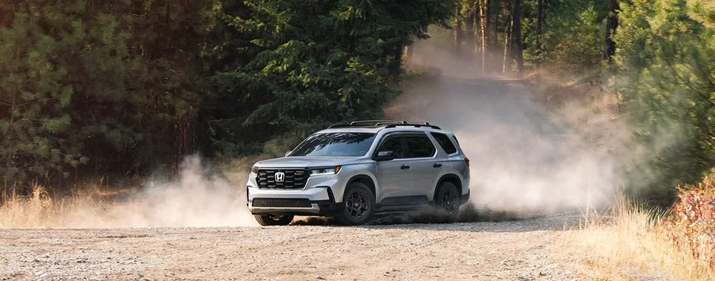 A silver 2024 Honda Pilot TrailSport is driving on a tree-lined trail.