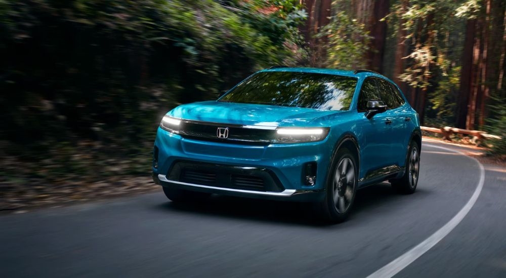 A teal 2024 Honda Prologue Elite is shown driving on a tree-line road.