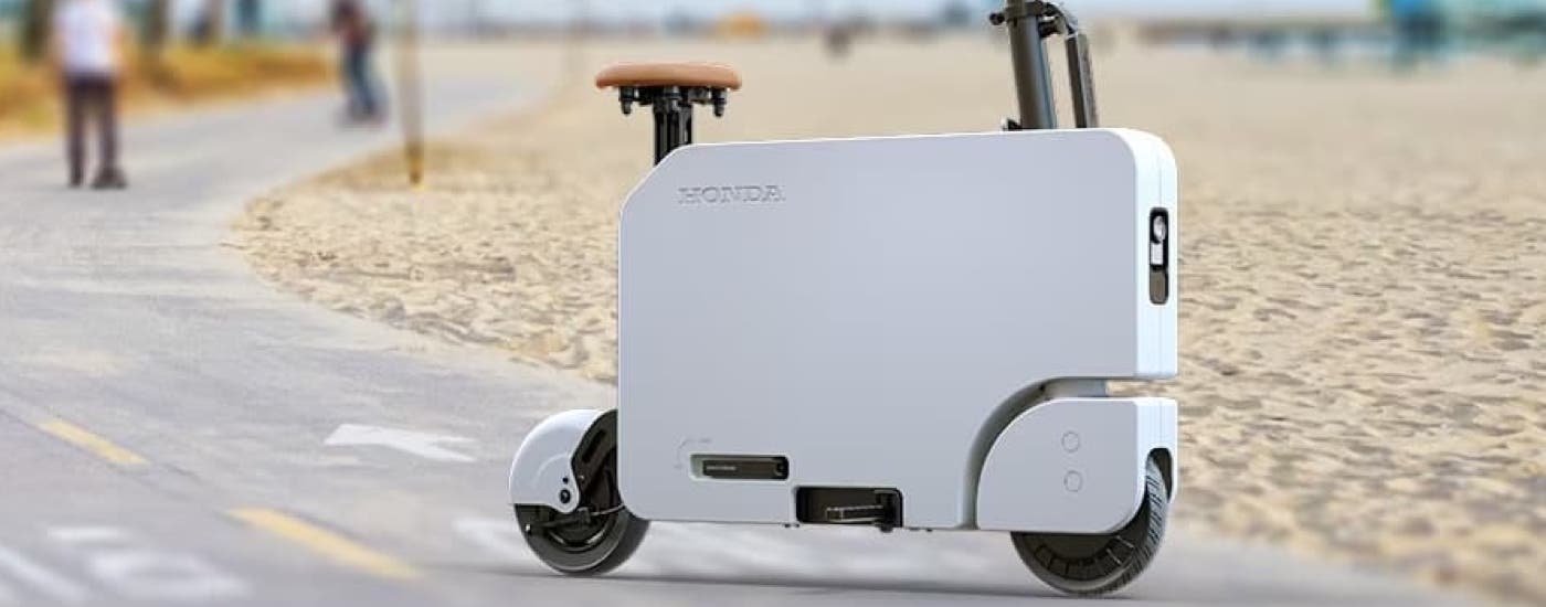 A white Honda Motocompacto is parked on a sidewalk next to a beach.