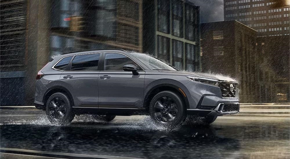 A grey 2023 Honda CR-V is shown from the side in the rain after leaving a dealer that has a Honda CR-V Hybrid for sale.