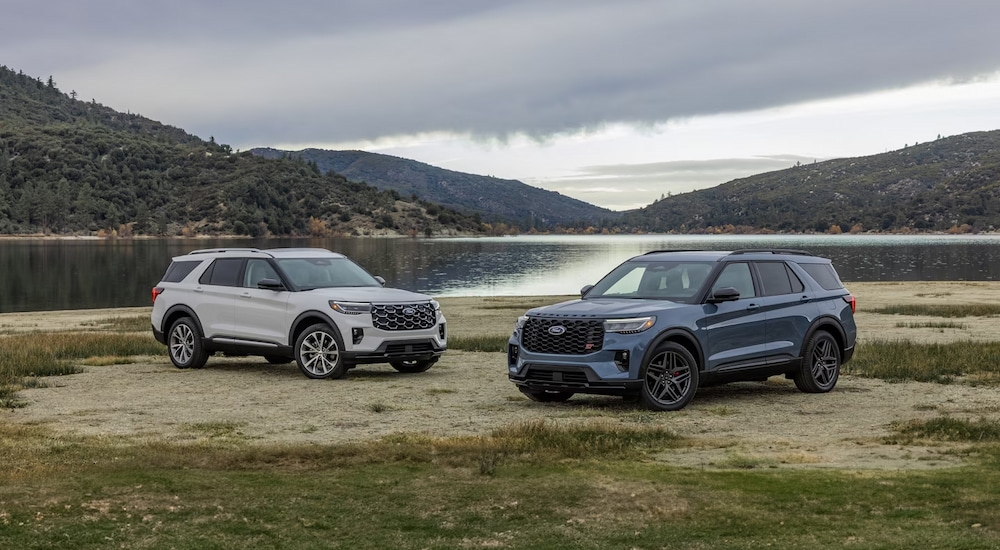 A white 2025 Ford Explorer Platinum and a blue 2025 Explorer ST parked at a lake.