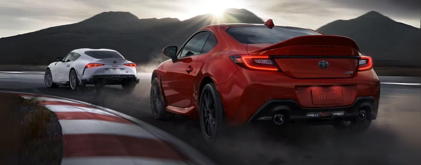 A white 2023 Toyota Supra and a red 2023 Toyota GR86 Premium are shown from the rear at an angle on a racetrack.