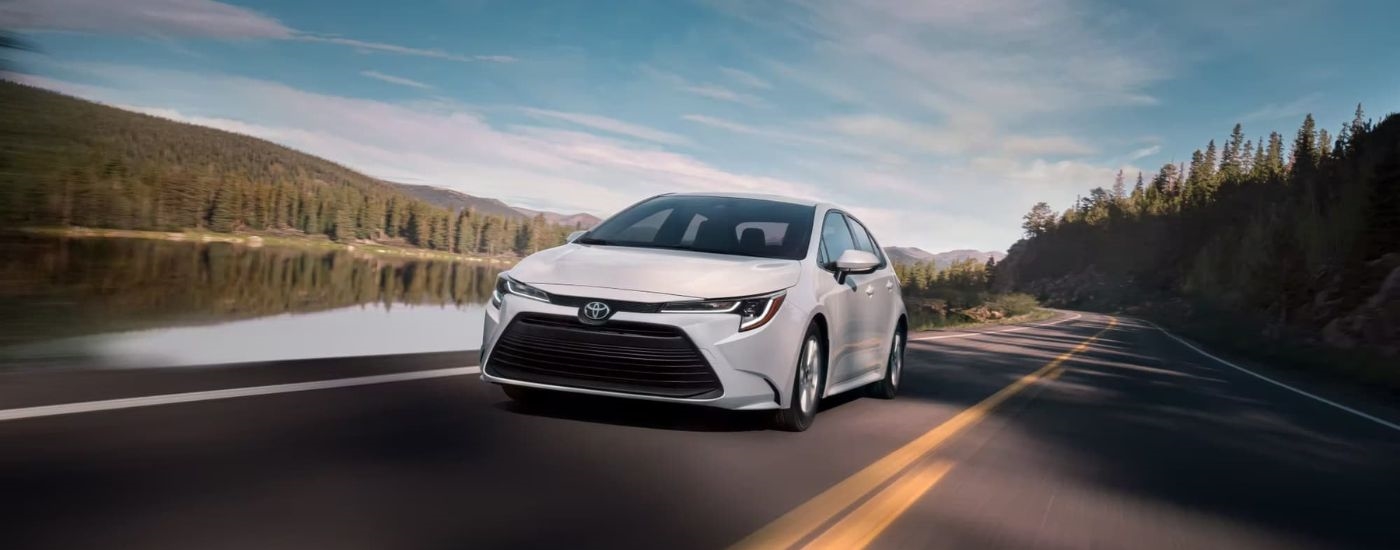 A white 2024 Toyota Corolla LE driving on a road past a body of water.