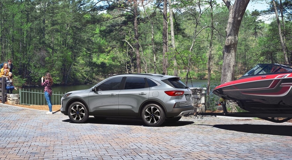 Side view of a silver 2025 Ford Escape towing a boat near water.