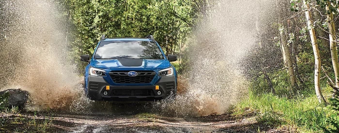 A front view of a blue 2024 Subaru Outback Wilderness driving over a muddy puddle.