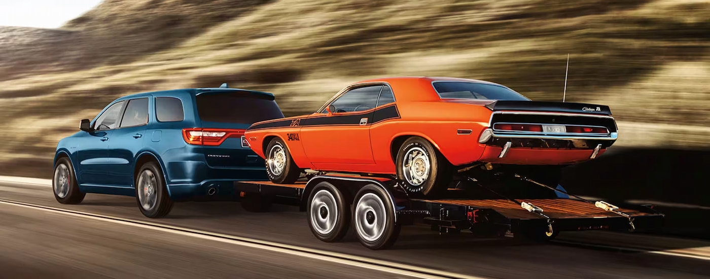 A blue 2024 Dodge Durango RT towing an orange 1970 Dodge Challenger T/A Six Pack on the highway.