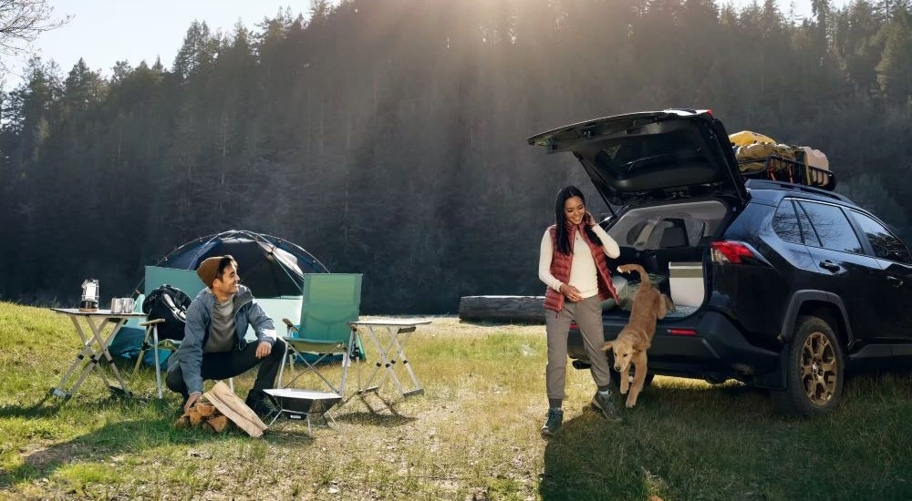 Two people are shown camping beside a black 2024 Toyota RAV4 Hybrid Woodland Edition parked off-road.