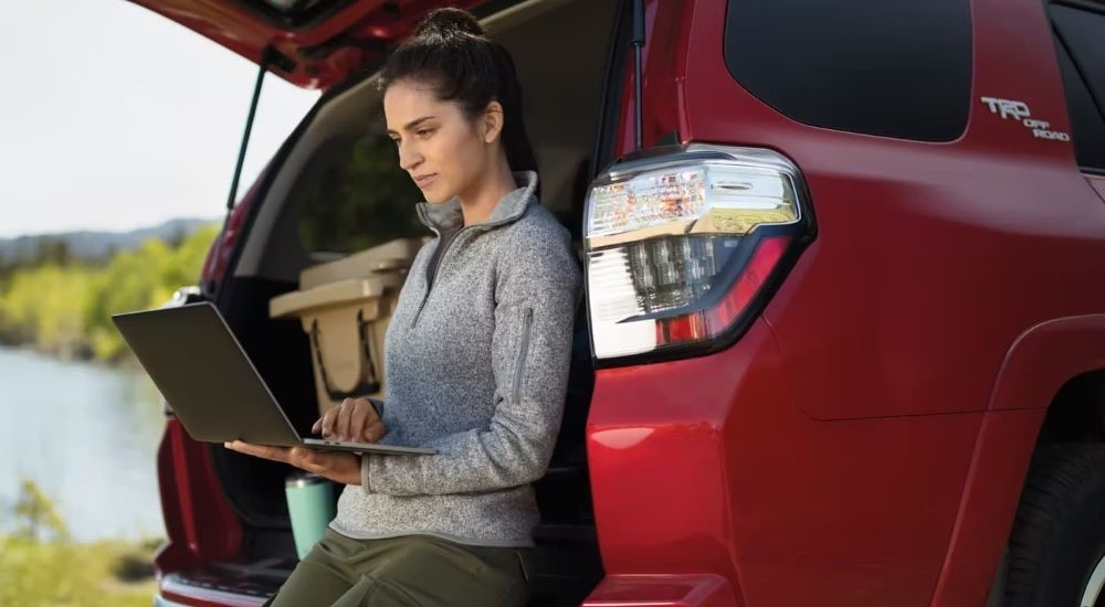 A woman using a laptop while leaning against the open hatchback of a red 2024 Toyota 4Runner TRD Off-Road.