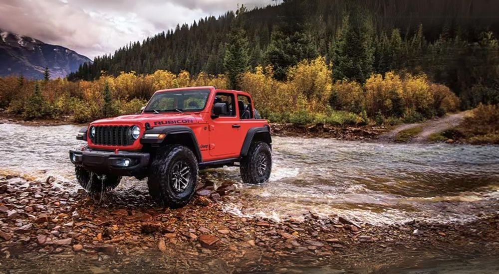 A red 2024 Jeep Wrangler Rubicon parked off-road by a shallow river.