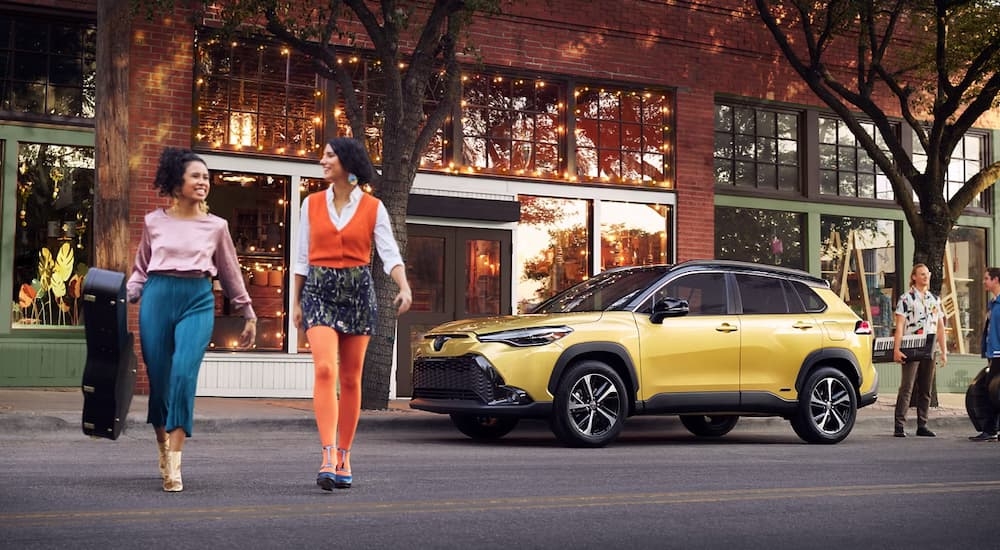 Two people walking from a yellow 2024 Toyota Corolla Cross Hybrid parked by a city curb.