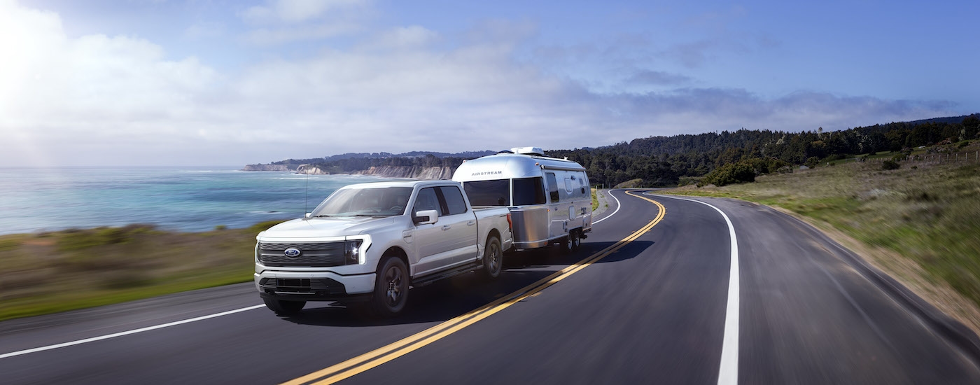 A white 2023 Ford F-150 Lightning is shown from the front at an angle while towing a camper.