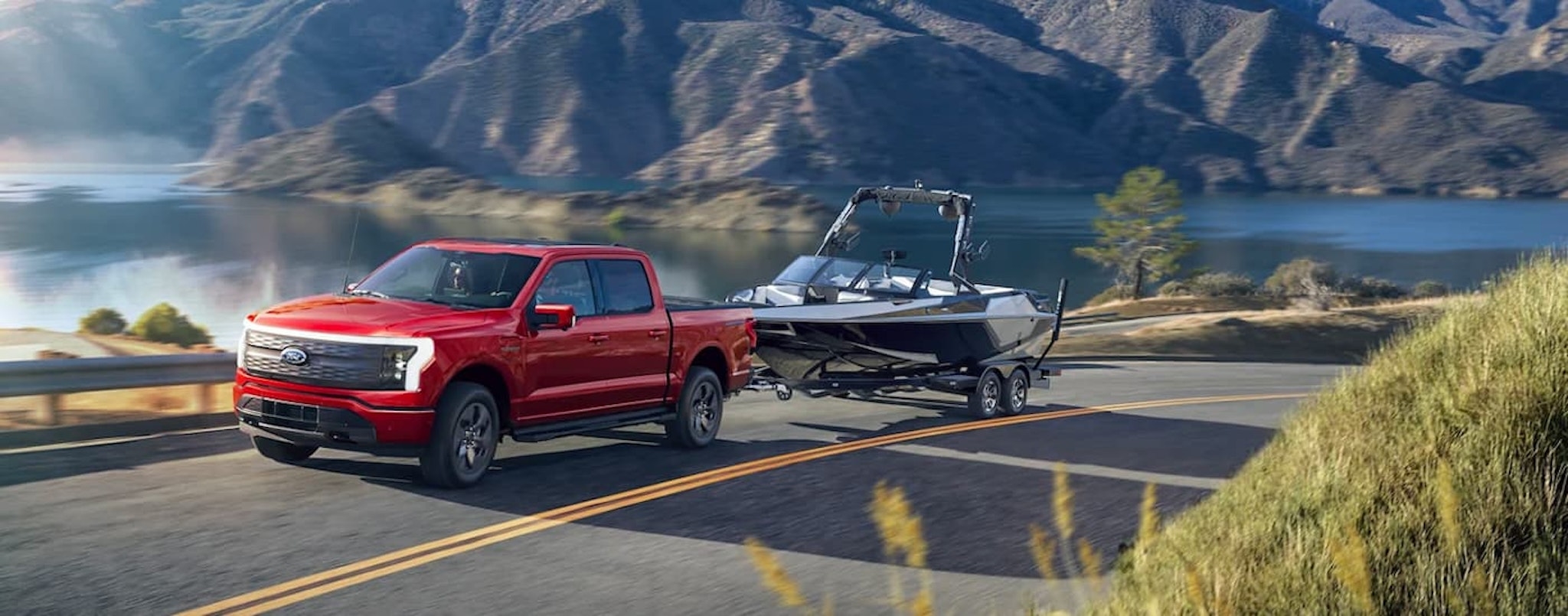 A red 2022 Ford F-150 Lightning is shown towing a boat past a lake after leaving a Ford dealer near you.