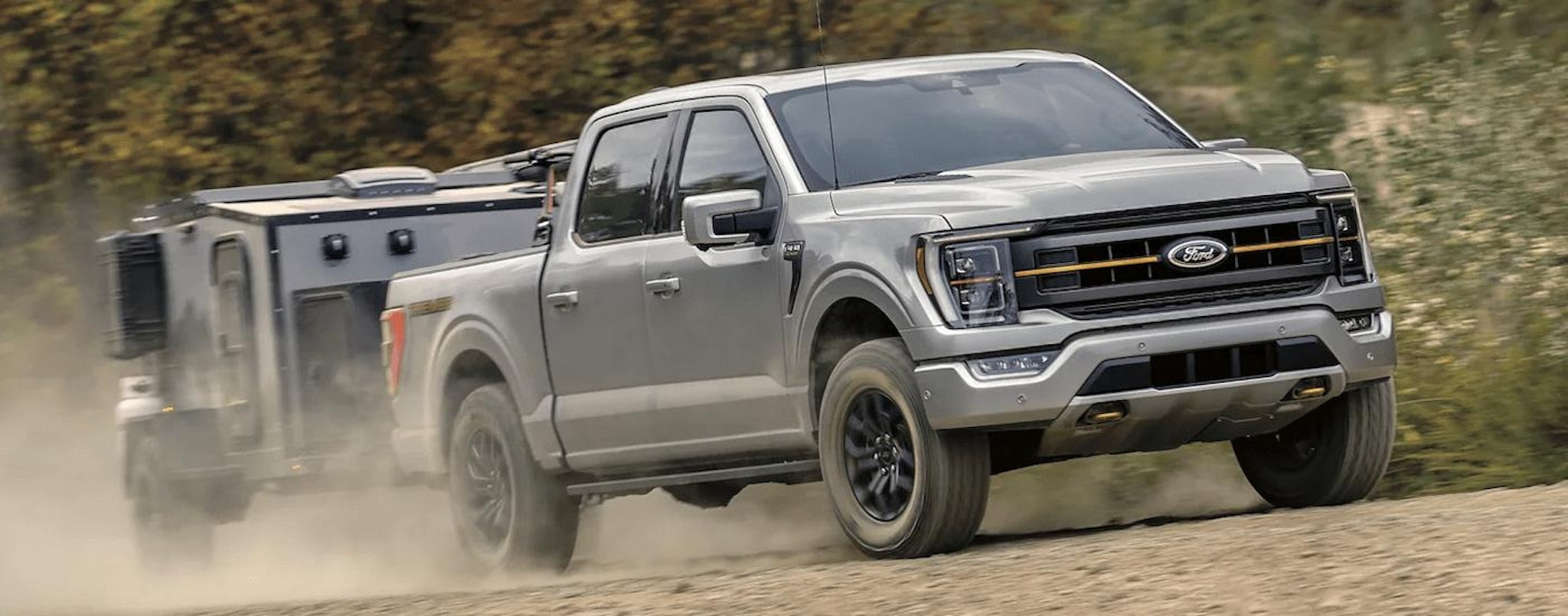 A grey 2023 Ford F-150 Tremor is shown towing a small camper on a dusty road.