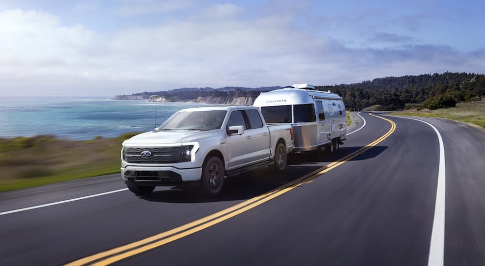 A white 2024 Ford F-150 Lightning is shown from the front at an angle while towing a camper.