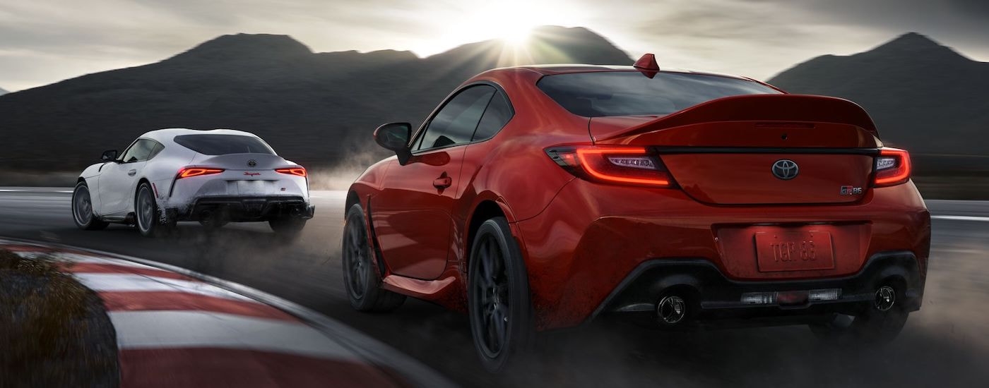 A red 2023 Toyota GR86 and a white 2023 Toyota Supra are shown from the rear on a racetrack.
