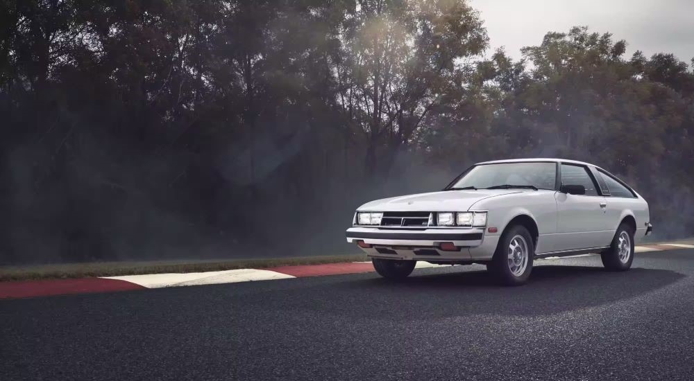 A white 1980 Toyota Supra is shown driving on a racetrack.