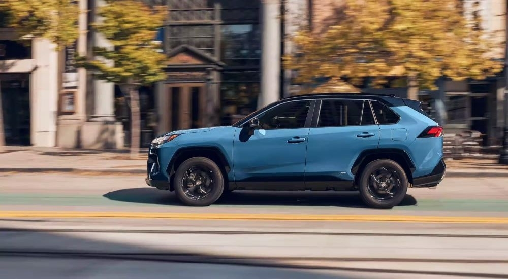 A blue 2023 Toyota RAV4 XSE Hybrid is shown from the side driving on a city street next to tall buildings.