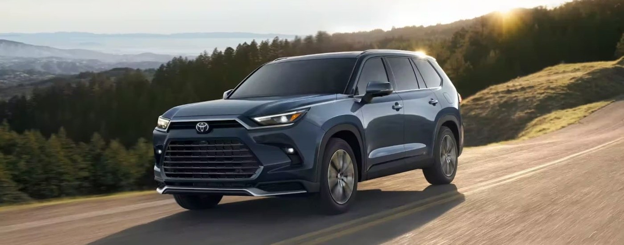 A blue 2024 Toyota Grand Highlander is shown driving on a sunny day.