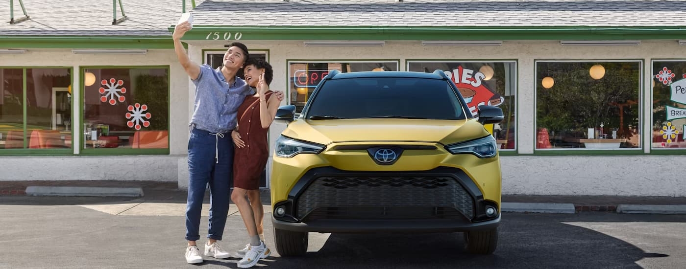 A yellow 2024 Toyota Corolla Cross Hybrid is parked outside of a store as a couple takes a selfie next to it.